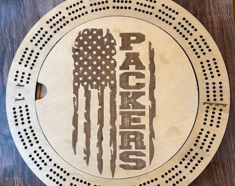 May include: A wooden cribbage board with a circular design. The board features a distressed American flag design with the word "PACKERS" in the center. The board has a light wood finish.
