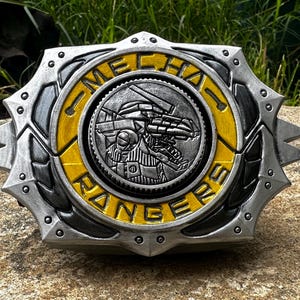 Puede incluir: Una insignia metálica detallada con las palabras "MECHA RANGERS" en letras amarillas. El centro presenta un diseño de robot plateado. La insignia tiene un borde plateado en forma de estrella con un anillo interior amarillo.