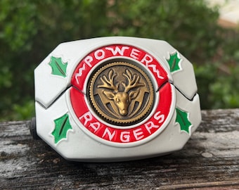May include: A white and silver Power Rangers belt buckle with a red and gold emblem. The emblem features a gold deer head and the words "POWER RANGERS". Green holly leaves decorate the edges.