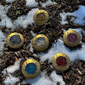 May include: Six round, gold-colored metal tokens with different colored centers. The tokens are arranged in a circle on a snowy, brown background.