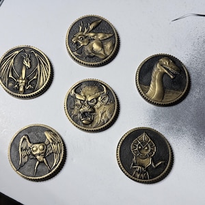 Puede incluir: Una colección de seis monedas de estilo antiguo con diseños en relieve detallados. Cada moneda presenta una criatura diferente: un dragón, un conejo, un monstruo, un dinosaurio, una criatura alada y una figura solar. Las monedas tienen una combinación de colores bronce y negro.