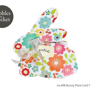 May include: A floral bunny place card template with the text "katie" written on it. The template is designed to be folded and has a ribbon attached to it. The template is part of the Pebbles in my Pocket collection and is numbered 498.