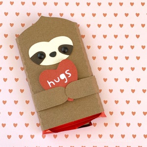 May include: A brown sloth holding a red heart with the word "hugs" on it. The sloth is made of paper and is holding a chocolate bar.