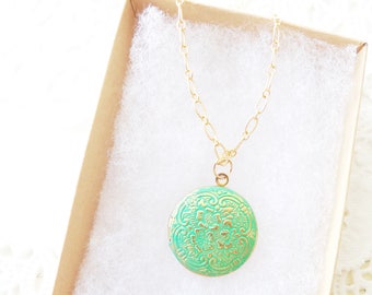 Puede incluir: Un collar de cadena dorada con un colgante redondo en forma de medallón. El medallón es verde con un diseño floral dorado.
