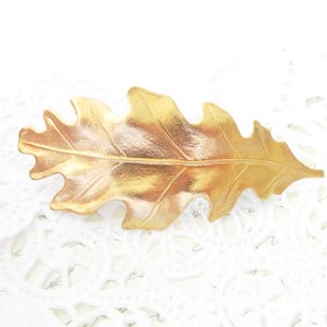 May include: A gold-colored leaf-shaped brooch with detailed veining. The brooch has a textured surface and is set against a white, lacy background. This nature-inspired accessory is suitable for adding a touch of elegance to any outfit.