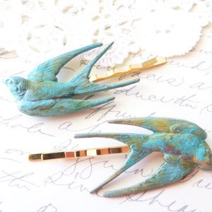 May include: Two turquoise blue metal swallow bird hair clips with gold metal barrettes. The birds have a distressed, aged look.