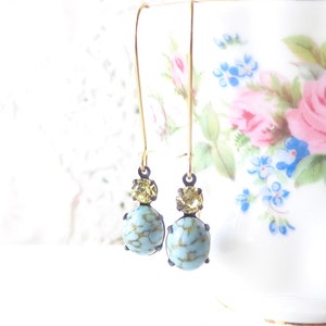 May include: A pair of dangle earrings with a light blue and gold design. The earrings feature a round, light blue stone with gold flecks and a smaller, round, gold stone above it.