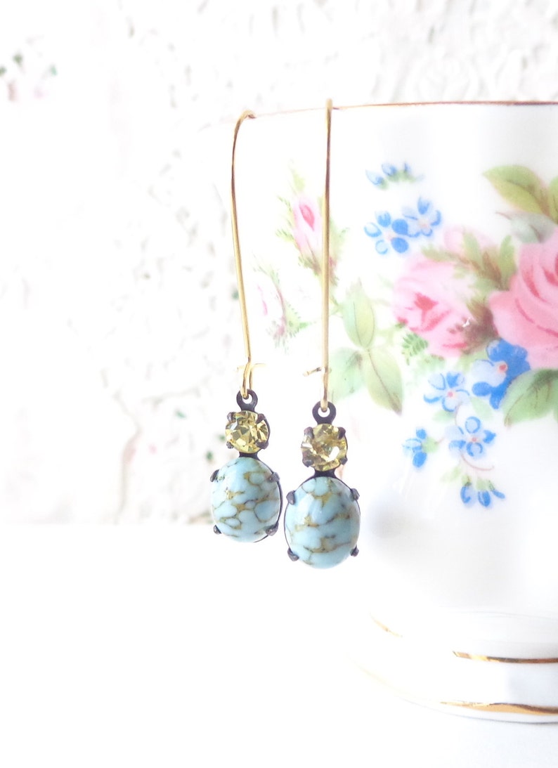 May include: A pair of dangle earrings with a light blue and gold design. The earrings feature a round, light blue stone with gold flecks and a smaller, round, gold stone above it.
