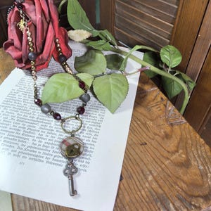 May include: A necklace featuring a chain of alternating matte grey, metallic gold, and deep red beads. A circular pendant with a plaid fabric inlay holds a small brass heart-shaped padlock and a dangling antique-style skeleton key charm.