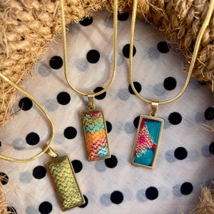 Puede incluir: Tres collares de cadena dorada con colgantes rectangulares. Cada colgante está hecho de tela con un patrón geométrico colorido diferente.