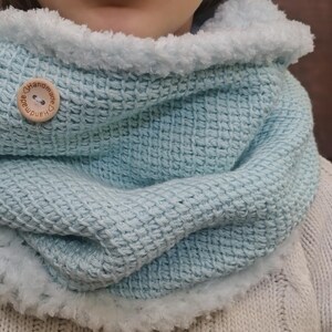 Könnte beinhalten: Ein hellblau gestrickter Cowl-Schal mit Waffelstruktur, verziert mit flauschigem weißem Material. Ein Holzknopf mit dem Text "handmade" sichert den Schal. Der Schal wird über einem cremefarbenen Pullover getragen.