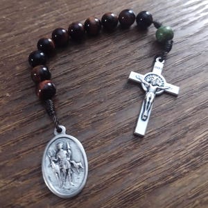 May include: A rosary with dark brown, spherical beads and a silver crucifix. The rosary also features a silver oval pendant with an engraved image of a saint. The beads are strung on a dark cord.