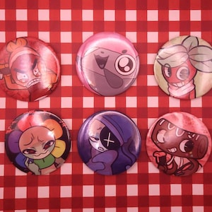 May include: A set of six circular pinback buttons featuring colorful cartoon character illustrations. The top row displays a red character, a pink creature, and a plant-like figure. The bottom row shows a multi-colored flower, a purple figure, and a brown character.