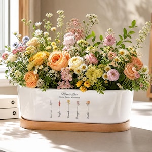Könnte beinhalten: Ein rechteckiger weißer Keramik-Blumentopf gefüllt mit einer Vielzahl bunter Blumen, darunter Rosen und Gänseblümchen. Der Topf steht auf einem Holzsockel und trägt den Text "Mom's Love Home Sweet Moments".