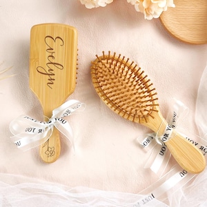 May include: Two wooden hairbrushes with natural bristles. One brush is rectangular with the name "Evelyn" engraved on it. Both brushes have white ribbons with floral designs and text. The brushes are on a light pink surface.