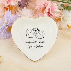 Può includere: Un portagioie in ceramica bianca a forma di cuore con bordo dorato. Il piatto presenta un'illustrazione nera di due lucchetti a forma di cuore ad incastro e il testo "August 20, 2025 Sophia & Jackson". Un anello d'oro con una pietra viola è in primo piano.