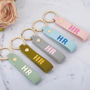 May include: Five leather keyrings in pastel shades of pink, light blue, grey, olive green, and light green. Each has the initials "HR" in a contrasting colour and a gold-coloured keyring. The keyrings are made of leather.