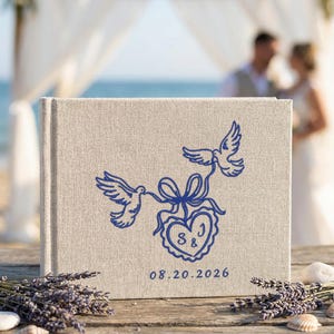 Puede incluir: Un álbum de fotos rectangular con cubierta de lino beige que presenta un diseño bordado en azul de dos palomas sosteniendo una cinta atada a un corazón. Dentro del corazón están las letras S y J, y la fecha 20.08.2026 está impresa debajo en azul.