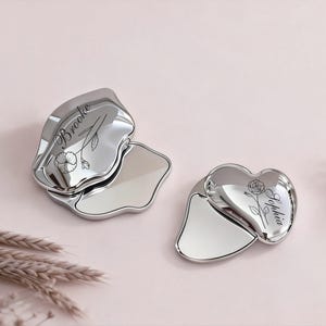 May include: Two silver-colored Gua Sha tools, one petal-shaped and one heart-shaped, are displayed on a pink surface. The petal-shaped tool has the name "Brooke" and a floral design. The heart-shaped tool has the name "Sophia" and a rose design.