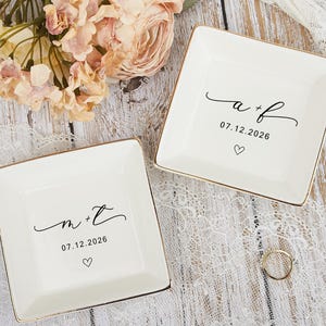May include: Two square, white ceramic ring dishes with gold trim. One dish has the initials "a + f", the date "07.12.2026", and a heart. The other dish has the initials "m + t", the date "07.12.2026", and a heart. Flowers and a ring are also visible.
