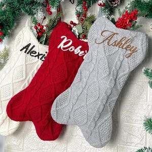 May include: Three cable-knit Christmas stockings in white, red, and gray. The white stocking features the name Alexis in black, the red stocking features the name Robert in white, and the gray stocking features the name Ashley in brown embroidery.