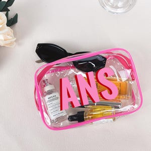 Op de afbeelding: Een doorzichtige, rechthoekige cosmetische tas met een roze rits en de letters "ANS" in roze. De tas bevat diverse schoonheidsproducten, waaronder een parfumfles, en is bedekt met een zwarte zonnebril.