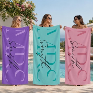 May include: Three rectangular beach towels in purple, teal, and pink. Each towel features large vertical block lettering spelling out a name, overlaid with a smaller, black cursive script text that reads Smith. The towels are held upright.