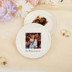 May include: Two white ceramic plates with gold rims, each featuring a printed photograph. One plate displays a photo of a mother and child, with the text "I love that you are my mom." The other plate has a photo of a mother and child. Gold rings with gemstones are also visible.