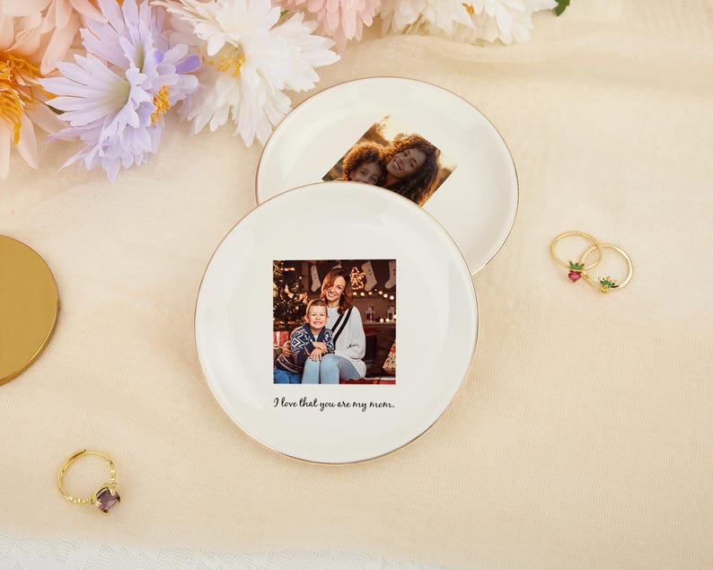May include: Two white ceramic plates with gold rims, each featuring a printed photograph. One plate displays a photo of a mother and child, with the text "I love that you are my mom." The other plate has a photo of a mother and child. Gold rings with gemstones are also visible.