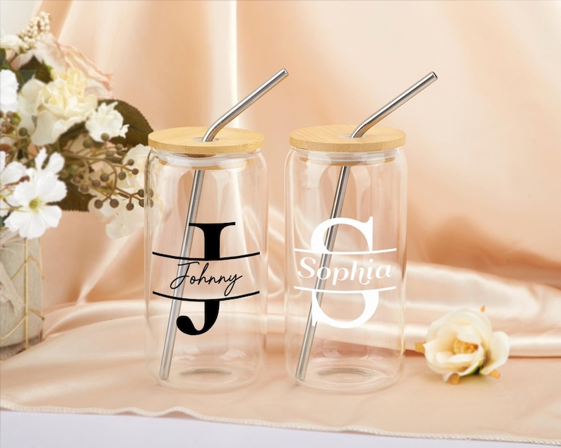Pode incluir: Dois copos de beber de vidro com tampas de bambu e canudos de a&ccedil;o inoxid&aacute;vel. Os copos t&ecirc;m nomes e iniciais personalizados, "Johnny" e "Sophia", impressos neles.