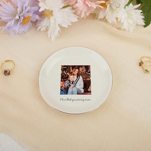 May include: A white ceramic trinket dish with a gold rim features a family photo and the text "I love that you are my mom." Two gold rings with gemstones and flowers are also visible, along with a gold ring with a purple gemstone.