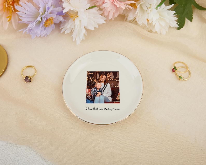 May include: A white ceramic trinket dish with a gold rim features a family photo and the text "I love that you are my mom." Two gold rings with gemstones and flowers are also visible, along with a gold ring with a purple gemstone.