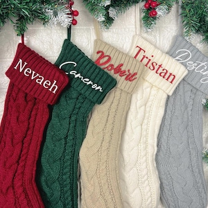 May include: Five cable-knit Christmas stockings in red, green, beige, white, and grey, each with a name embroidered on the cuff: Nevaeh, Cameron, Robert, Tristan, and Destiny. The stockings are hanging against a white brick wall, decorated with evergreen branches.