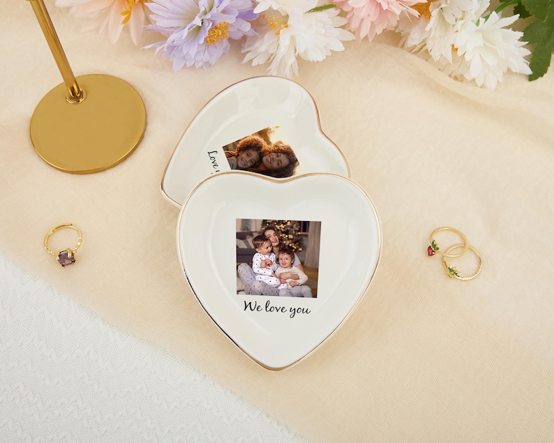 May include: Two heart-shaped, white ceramic trinket dishes with gold trim, each holding a photograph. One dish has the text "We love you". A gold ring with a purple stone and two gold rings with floral details are also present.