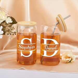 Pode incluir: Dois copos de beber de vidro transparente com tampas de bambu e canudos. Os copos est&atilde;o cheios de um l&iacute;quido castanho claro. Os copos t&ecirc;m letras brancas que dizem "Sophia" e "Daniel".