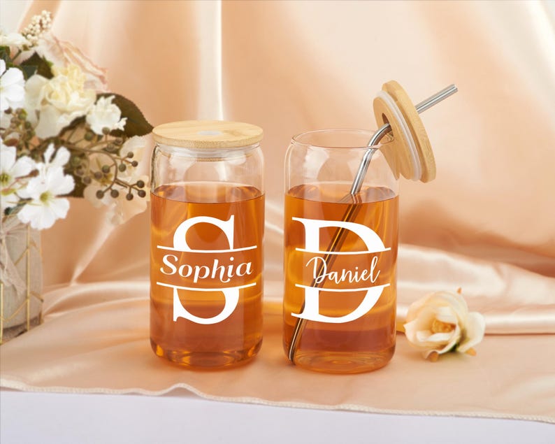 Pode incluir: Dois copos de beber de vidro transparente com tampas de bambu e canudos. Os copos est&atilde;o cheios de um l&iacute;quido castanho claro. Os copos t&ecirc;m letras brancas que dizem "Sophia" e "Daniel".