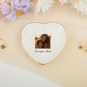 May include: A heart-shaped, white ceramic trinket dish with a gold rim, featuring a printed photo of a mother and child. The dish has the text "Love you, Mom". A gold ring with a purple stone and two gold rings with floral accents are also visible.