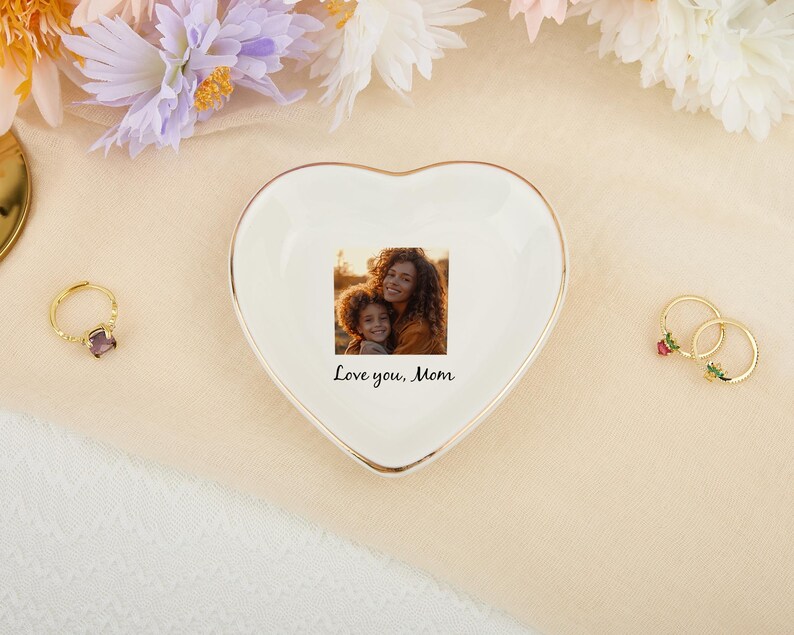 May include: A heart-shaped, white ceramic trinket dish with a gold rim, featuring a printed photo of a mother and child. The dish has the text "Love you, Mom". A gold ring with a purple stone and two gold rings with floral accents are also visible.