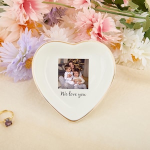 May include: A heart-shaped, off-white ceramic trinket dish with a gold rim. The dish features a small photo and the text "We love you". The background includes flowers and a gold ring.