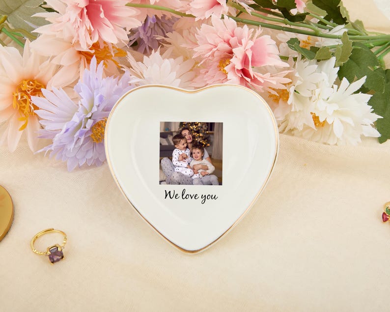 May include: A heart-shaped, off-white ceramic trinket dish with a gold rim. The dish features a small photo and the text "We love you". The background includes flowers and a gold ring.