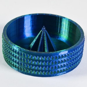 May include: A blue and green 3D printed dish with a central cone-shaped divider. The dish has a textured outer rim.