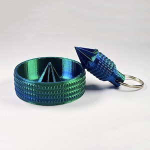 May include: A green and blue 3D printed keychain with a small, pointed, cone-shaped top and a circular base. The keychain has a textured surface and a key ring.
