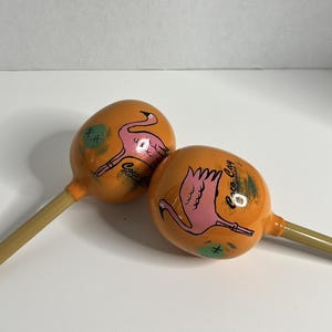 Könnte beinhalten: Zwei orangefarbene Maracas mit Flamingo-Illustrationen. Jede Maraca hat einen Holzgriff und einen runden Kopf. Der Flamingo ist rosa und die Maracas haben grüne Akzente. Der Text "Coco Cay Bahamas" ist sichtbar.