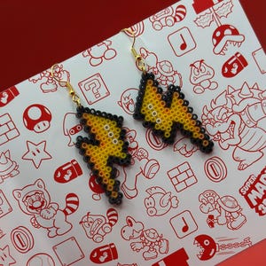 May include: A pair of yellow and black pixelated lightning bolt earrings. The earrings are made of perler beads and have gold hooks.