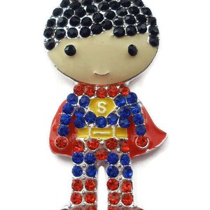 May include: A silver charm featuring a cartoon superhero with black hair, a red and blue suit, and a yellow cape. The charm is decorated with red, blue, and black rhinestones.