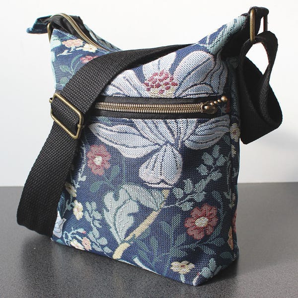 May include: A crossbody bag featuring a dark blue woven fabric with a floral pattern in shades of light blue, white, and muted red. The bag includes a black adjustable strap, a top zip closure, and a front exterior pocket with a brass-toned zip.