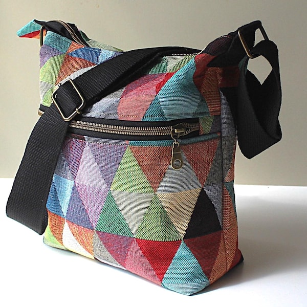 May include: A crossbody bag featuring a vibrant, multi-coloured geometric triangle pattern in shades of red, blue, green, yellow, and purple. The bag includes a black adjustable shoulder strap, a top zip closure, and a front horizontal zip pocket.