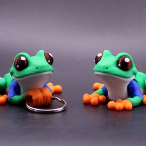 May include: Two handmade frog figurines with large, expressive eyes. The frogs are primarily green with blue accents and orange feet. One frog holds a keychain ring. The figurines are likely made of polymer clay or a similar material.