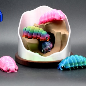 May include: A 3D printed display featuring a white arched structure with a brown base, housing three small segmented creatures in pink, rainbow, and dark blue. Two additional segmented creatures, one pink and one teal, sit in front of the display.