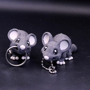 Puede incluir: Dos llaveros con forma de ratón gris con orejas y ojos blancos. Un ratón sostiene un llavero de anillo plateado y el otro tiene un llavero de cadena plateada.